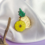 A handcrafted Pineapple Pin accessory with green leaves and a yellow ring rests on white satin fabric, beside textured brown driftwood on a purple surface.