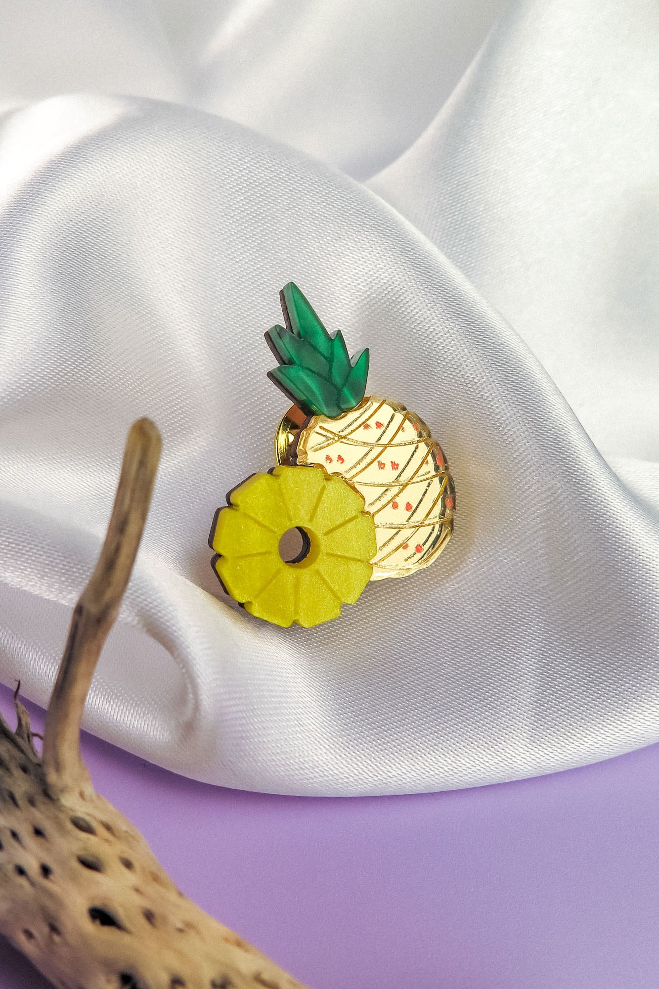 A handcrafted Pineapple Pin accessory with green leaves and a yellow ring rests on white satin fabric, beside textured brown driftwood on a purple surface.