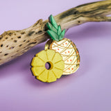 A decorative handcrafted Pineapple Pin with green leaves and a sliced section rests on a piece of driftwood against a pastel purple background.