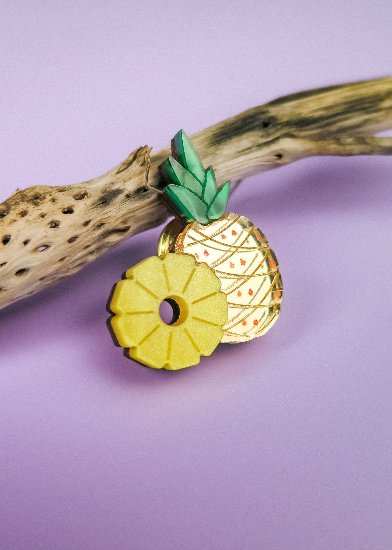 A decorative handcrafted Pineapple Pin with green leaves and a sliced section rests on a piece of driftwood against a pastel purple background.