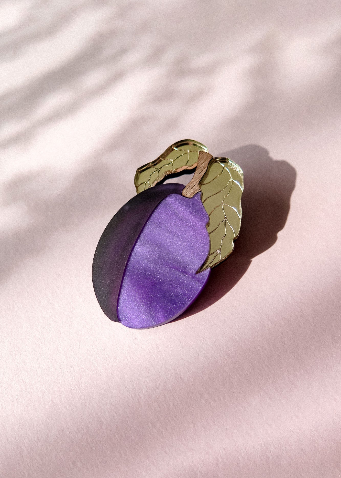 A decorative brooch shaped like a plum with two green leaves, crafted from acrylic and recycled wood, rests on a pale pink surface with soft shadows.