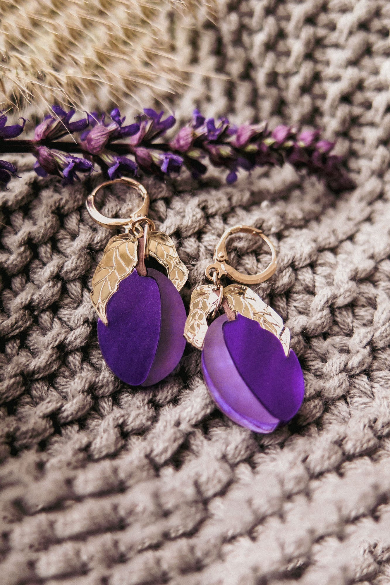 Pair of gold and purple plum earrings with textured gold accents, displayed on a knitted beige fabric with a sprig of purple flowers alongside. These handcrafted earrings add a touch of elegance to any outfit.
