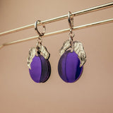 A pair of elegant purple plum earrings with silver leaf accents hang from a gold-colored rod against a beige background.