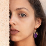 A woman with long brown hair and elegant purple plum earrings looks directly at the camera, standing next to a textured beige wall, with soft natural light highlighting her face.