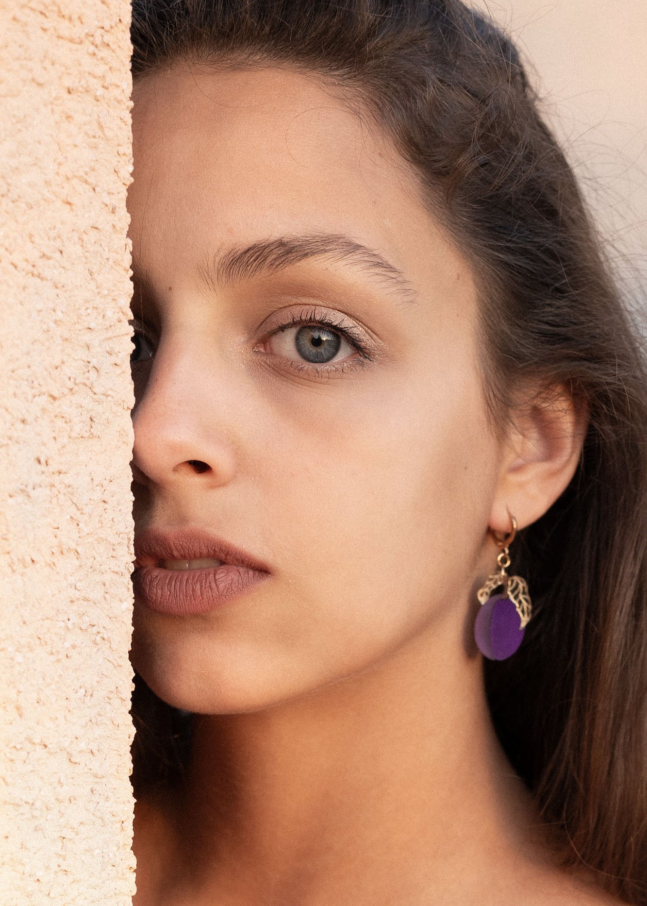 A woman with long brown hair and elegant purple plum earrings looks directly at the camera, standing next to a textured beige wall, with soft natural light highlighting her face.