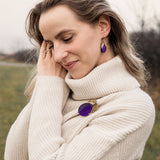 A woman wearing a cream-colored turtleneck sweater stands outdoors, smiling gently with eyes closed. She has wavy hair and wears matching purple jewelry—a handcrafted brooch and earrings. The grassy background is slightly blurred.