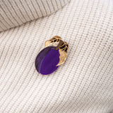 A handcrafted brooch shaped like a gold and purple eggplant with two gold leaves is pinned to a cream-colored ribbed sweater.