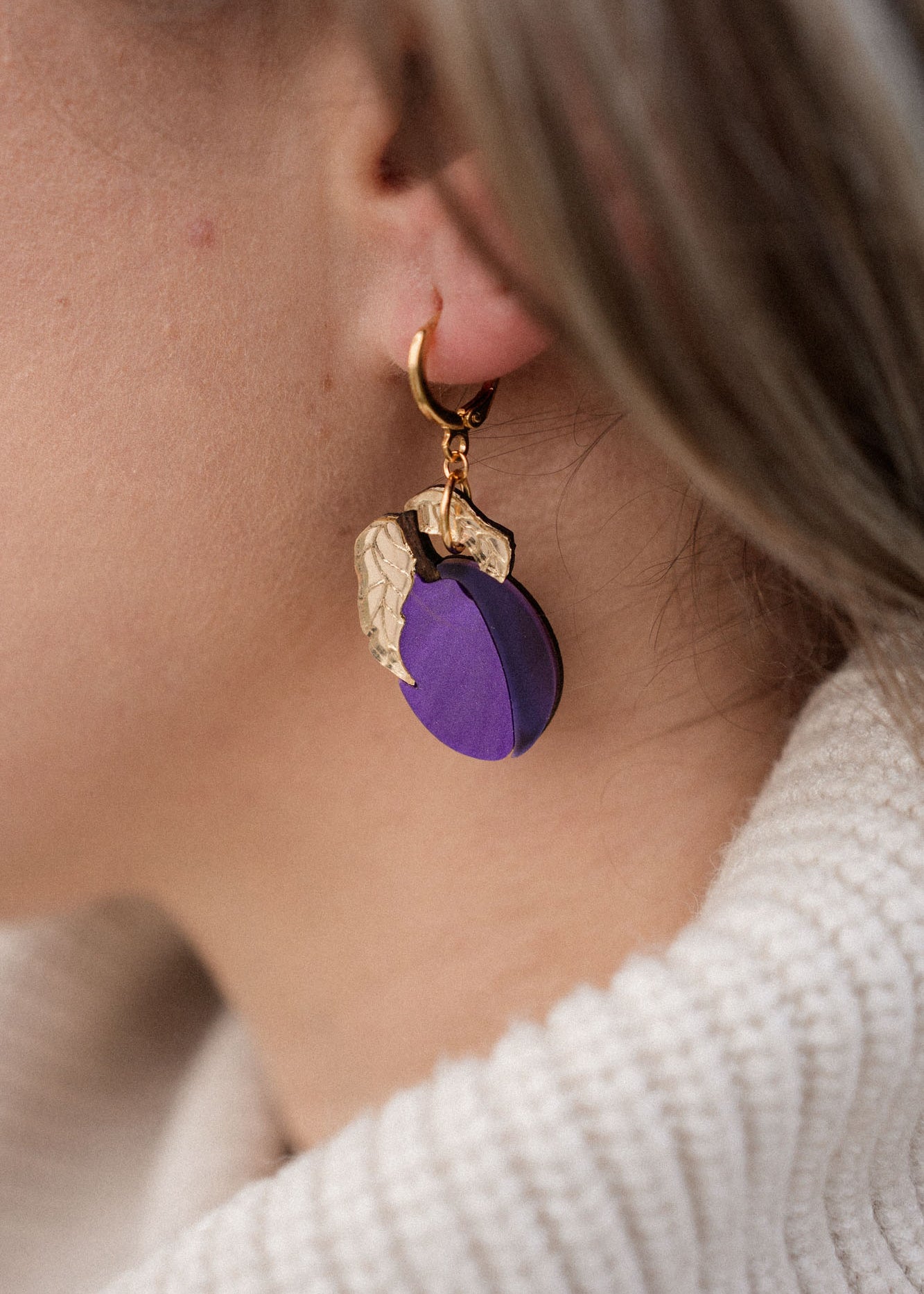 Close-up of a person wearing elegant purple plum earrings shaped like fruit with gold leaves. The person has light brown hair and is wearing a cream-colored, ribbed turtleneck sweater.