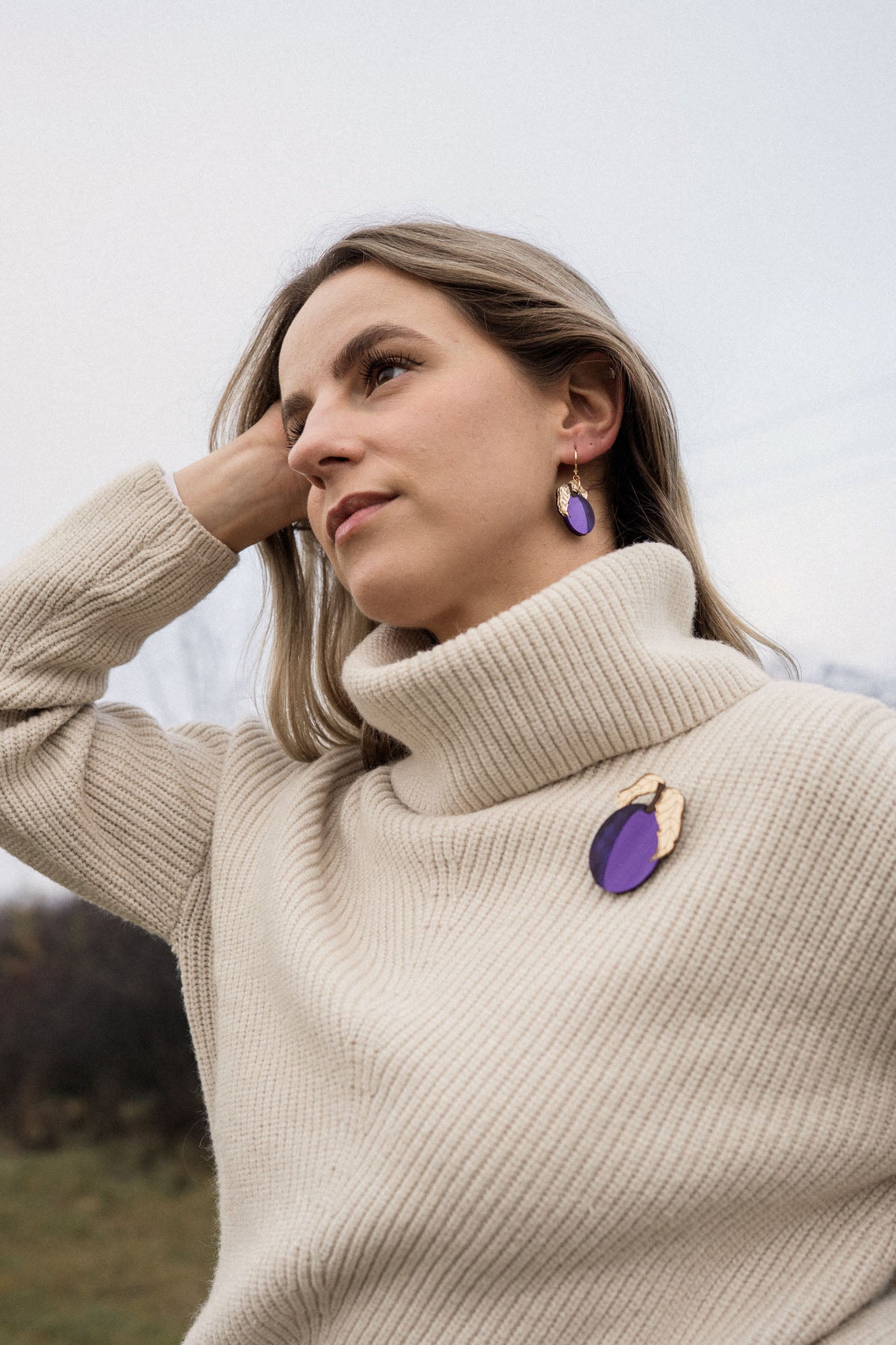 A woman with blonde hair wearing a cream-colored turtleneck sweater, handcrafted purple plum earrings, and a matching purple brooch stands outdoors, looking thoughtful with her hand resting on her head.