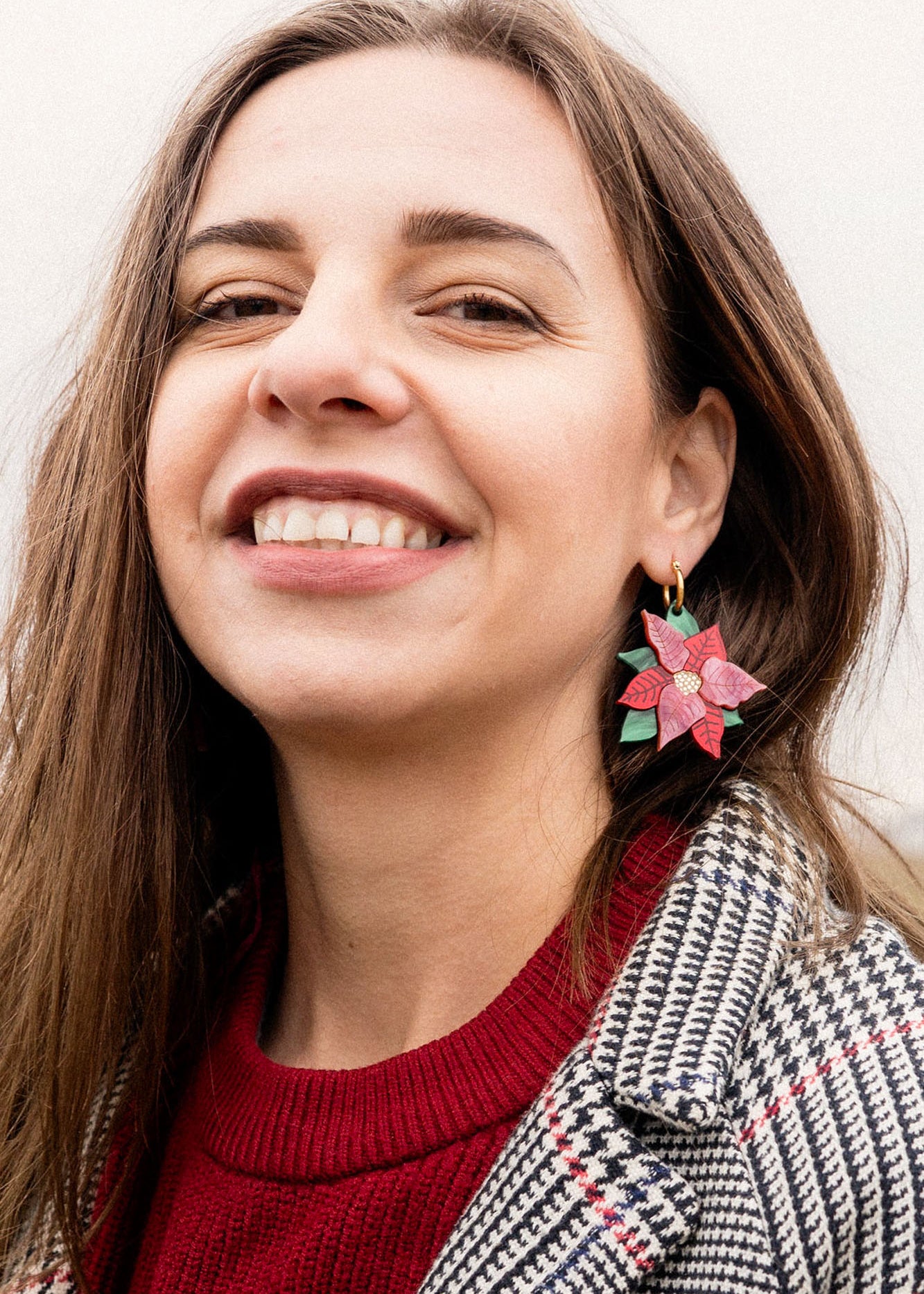 A woman with long brown hair smiles, wearing a red sweater and checkered coat. She showcases festive jewelry with large Poinsettia Statement Hoops, holiday earrings shaped like red and green poinsettias. The background is softly blurred.