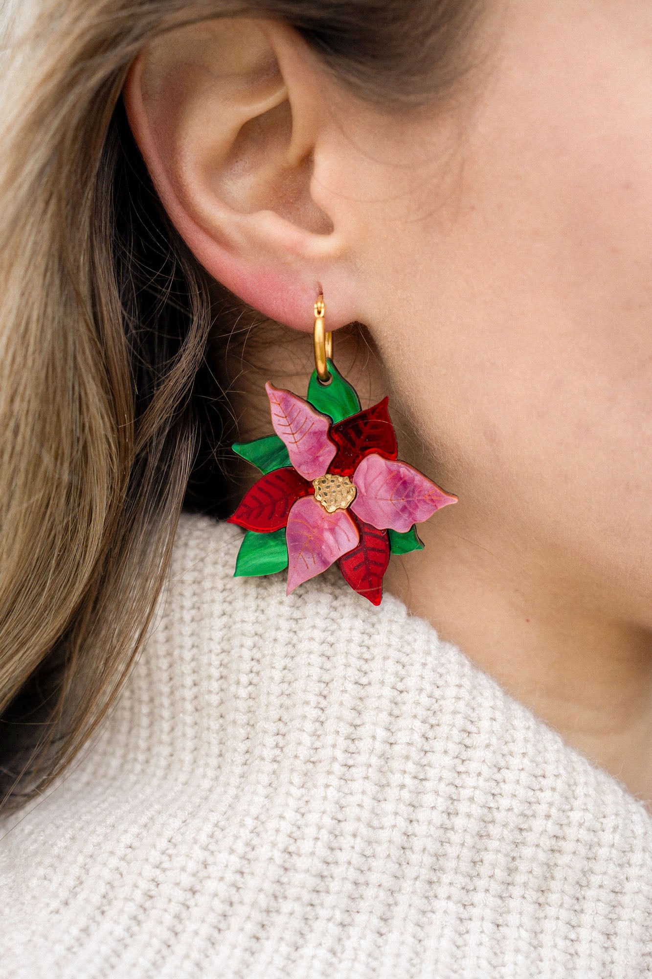 Close-up of a person's ear wearing festive jewelry: colorful, flower-shaped Poinsettia Statement Hoops with red and pink petals, green leaves, and a gold center, paired with a cream knit sweater.