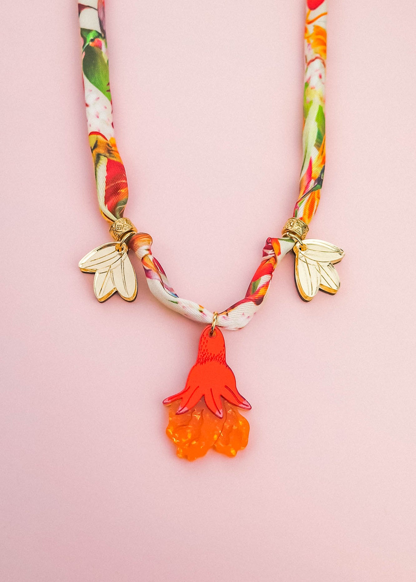 A colorful handmade necklace with a floral-patterned fabric band, two gold flower charms, and a central pendant shaped like an orange and red flower—perfect as a unique pomegranate flower necklace—displayed on a light pink background.