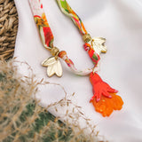A colorful silk cord necklace with gold leaf charms and an orange-red hand-shaped pendant, displayed on white fabric with a woven basket and beige dried grass nearby.