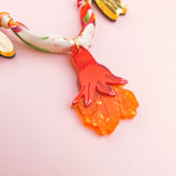 A colorful handmade necklace with a twisted fabric band and gold leaves features an orange, hand-shaped pendant with textured details, set against a light pink background.