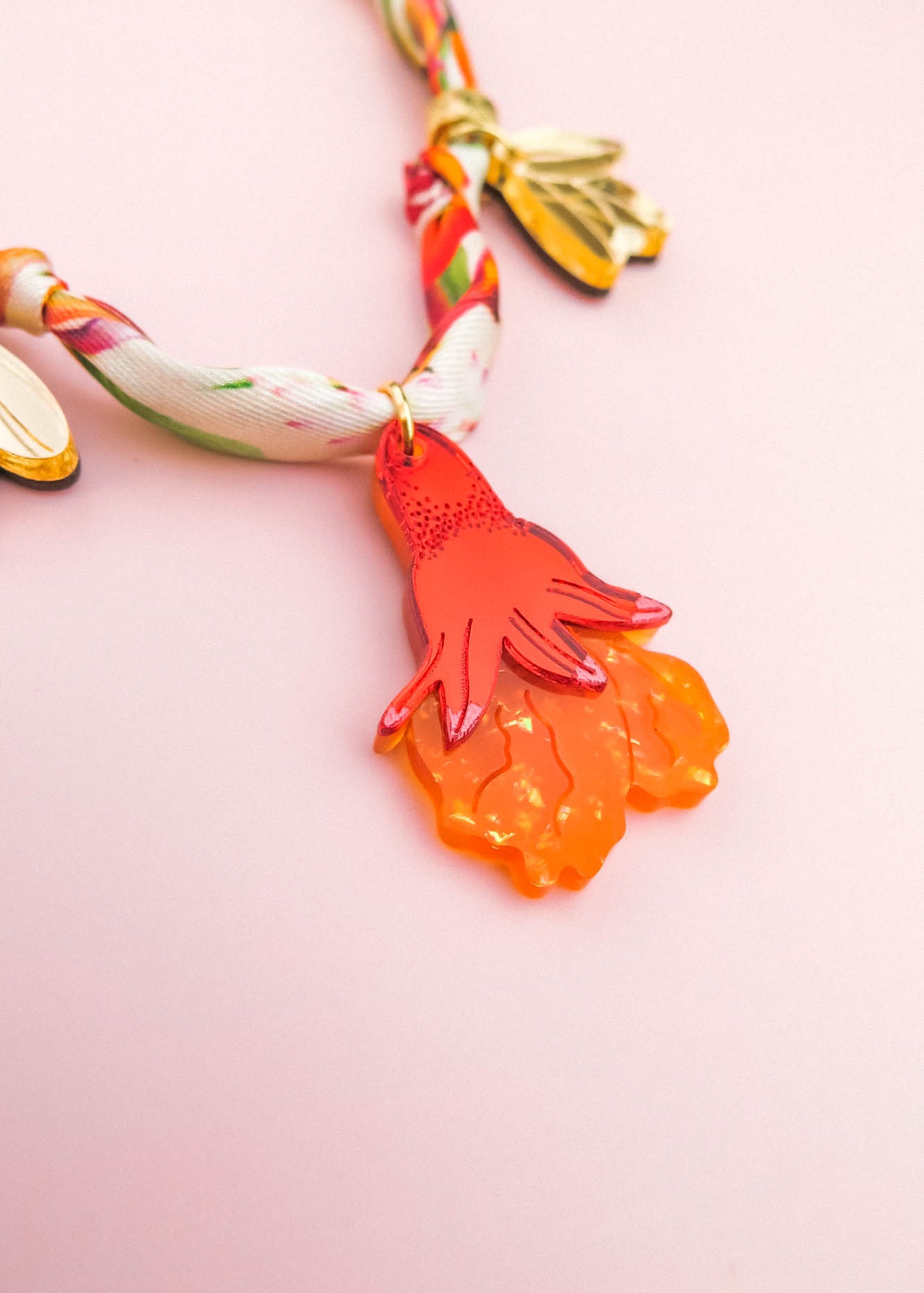A colorful handmade necklace with a twisted fabric band and gold leaves features an orange, hand-shaped pendant with textured details, set against a light pink background.