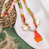 A colorful handmade necklace with floral patterns, featuring two gold and white wing-shaped charms and a bright orange hand-shaped pendant, is displayed on a white cloth and green textured background.