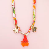 A colorful silk cord necklace with a floral pattern, featuring two gold flower charms and a red-orange pomegranate flower pendant, displayed on a pale pink background.
