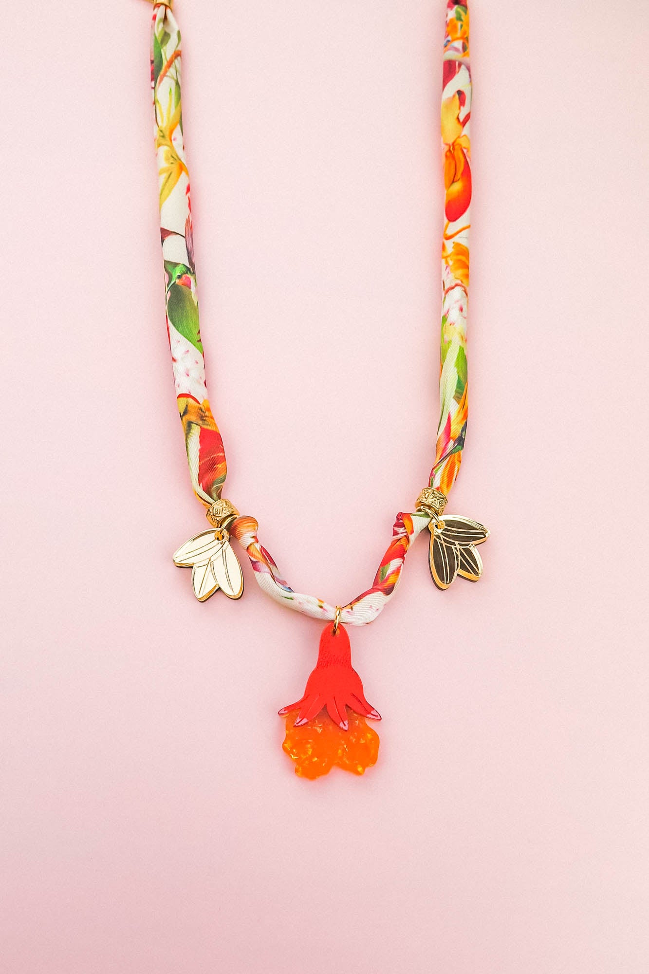 A colorful silk cord necklace with a floral pattern, featuring two gold flower charms and a red-orange pomegranate flower pendant, displayed on a pale pink background.