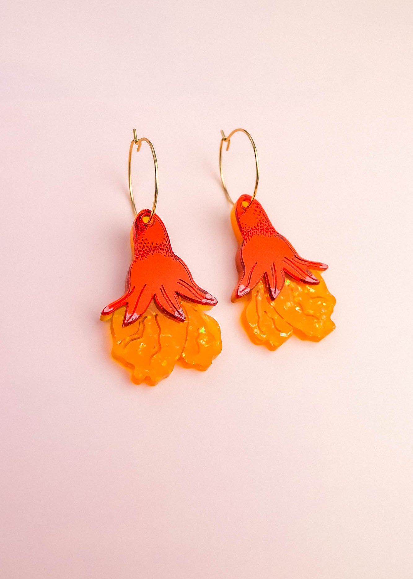 A pair of Little Turtle Hoop Earrings featuring gold hoops and pendants shaped like orange and yellow hand-drawn flowers, set against a pale pink background—beautifully handcrafted earrings for a playful, artistic touch.