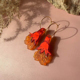 A pair of handcrafted earrings with orange and red hand-shaped charms and translucent, wavy orange accents dangle from gold hoops, resting on a pale pink surface near a rock and dried plant shadows.