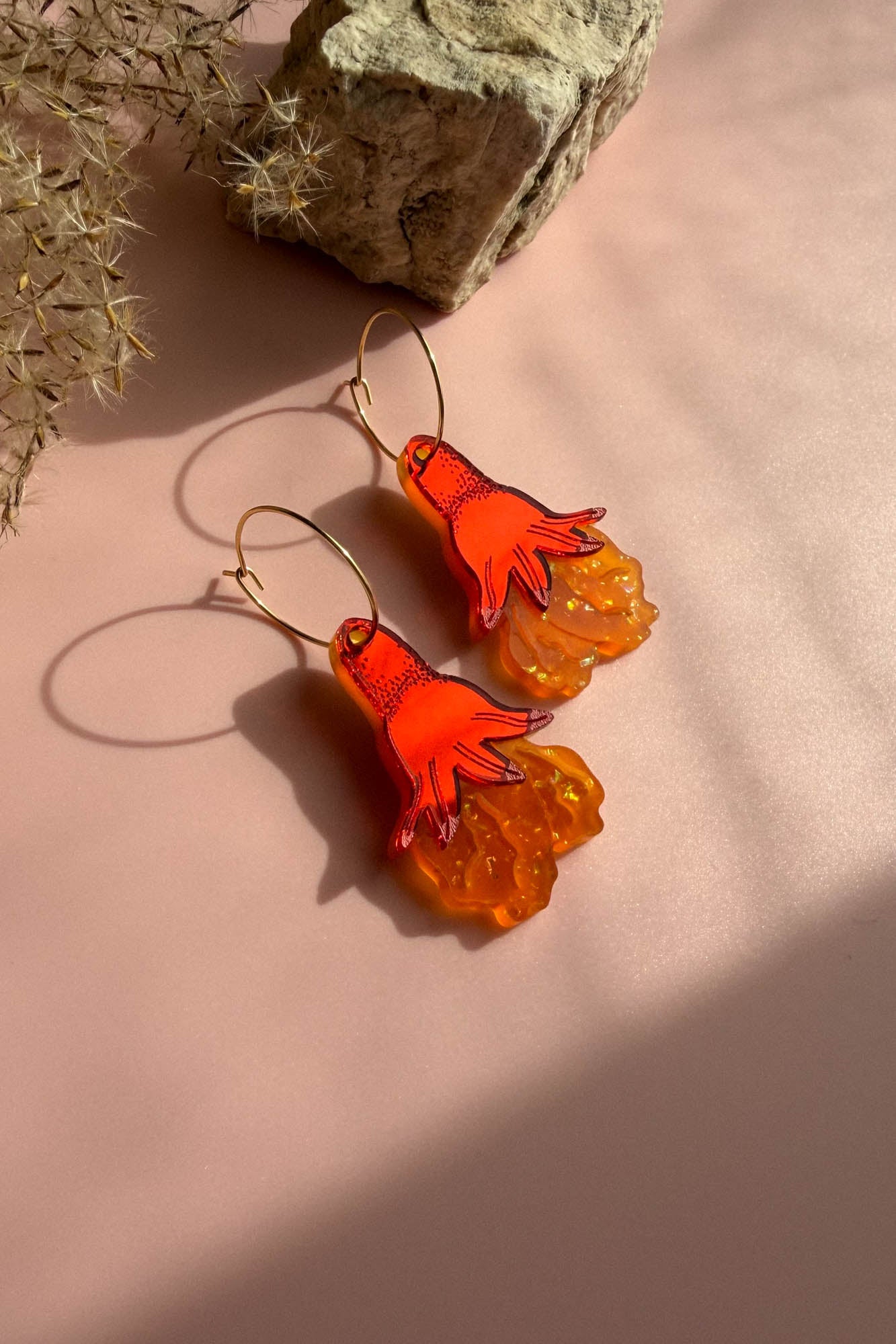 A pair of handcrafted earrings with orange hand-shaped charms and textured translucent orange bases dangle from gold hoops, displayed on a pink surface near a rock and dried plant.