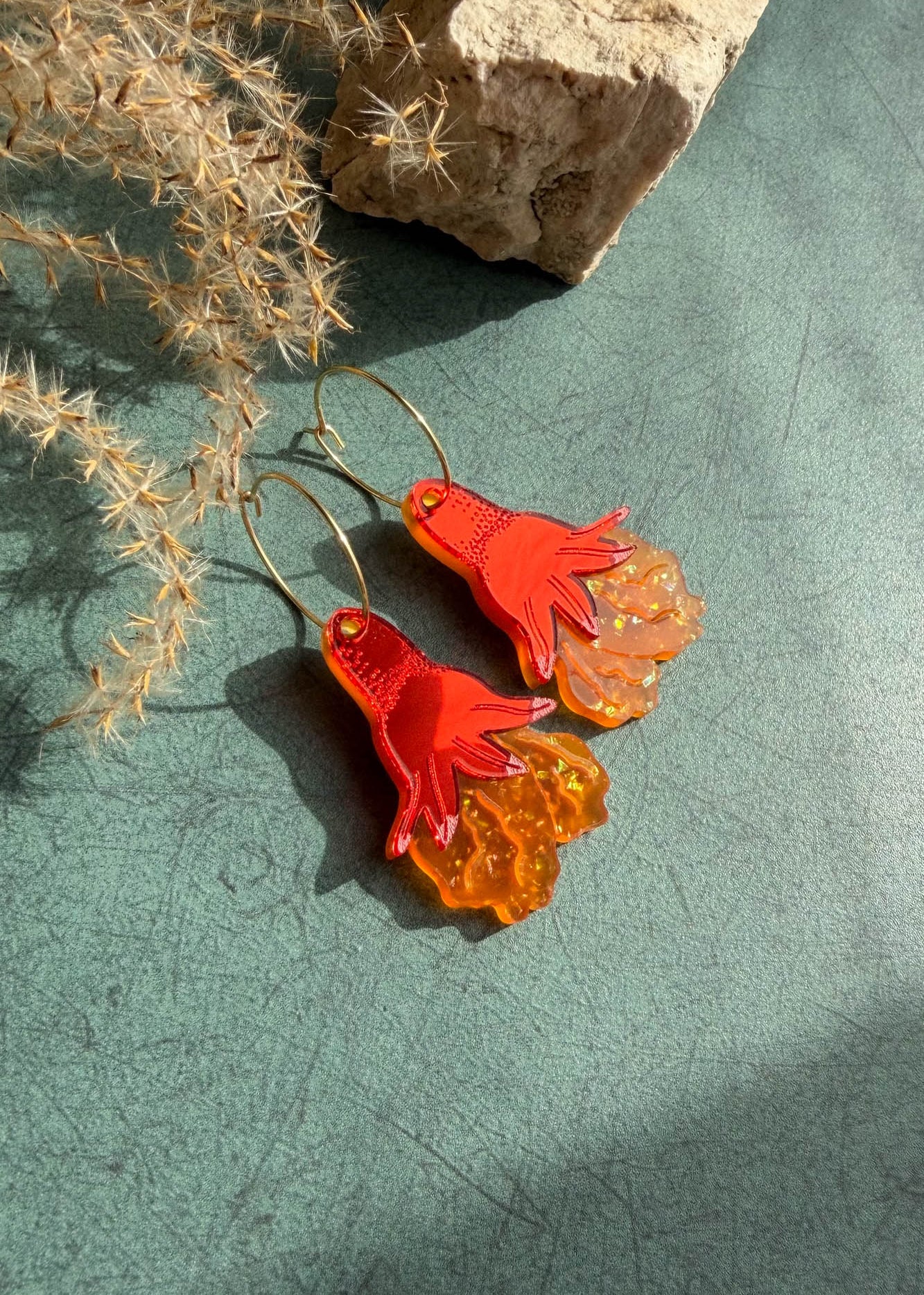 A pair of handcrafted earrings shaped like bright red-orange octopus tentacles with wavy, translucent lower sections, lying on a green surface near dried grass and a beige rock.