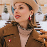 A woman in a tan beret and brown coat stands outside on a snowy day, wearing bold, handcrafted earrings with a bird design, a matching pin, tan gloves, and red lipstick. Snowflakes sprinkle her hair and coat.
