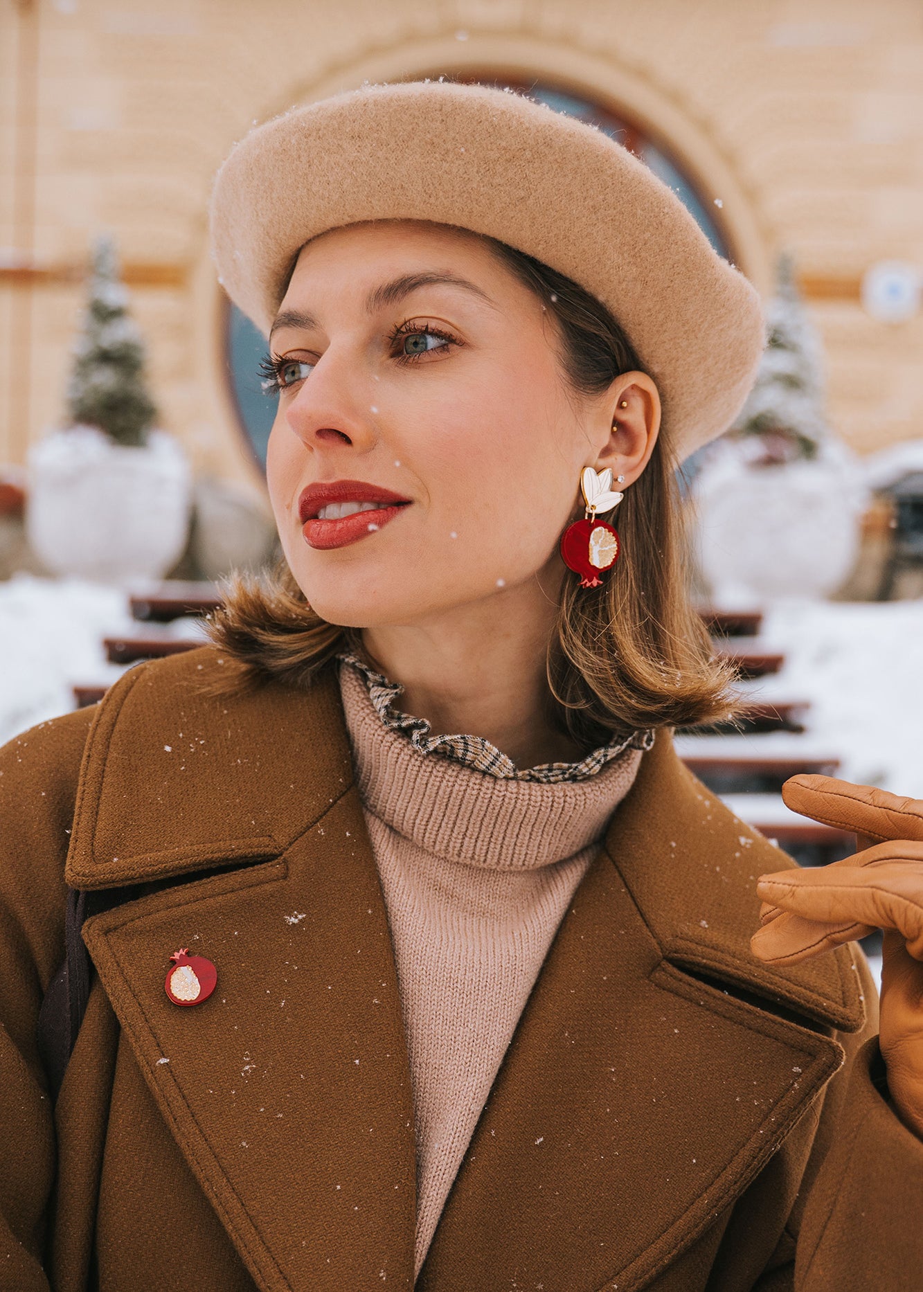 A woman in a tan beret and brown coat stands outside on a snowy day, wearing bold, handcrafted earrings with a bird design, a matching pin, tan gloves, and red lipstick. Snowflakes sprinkle her hair and coat.