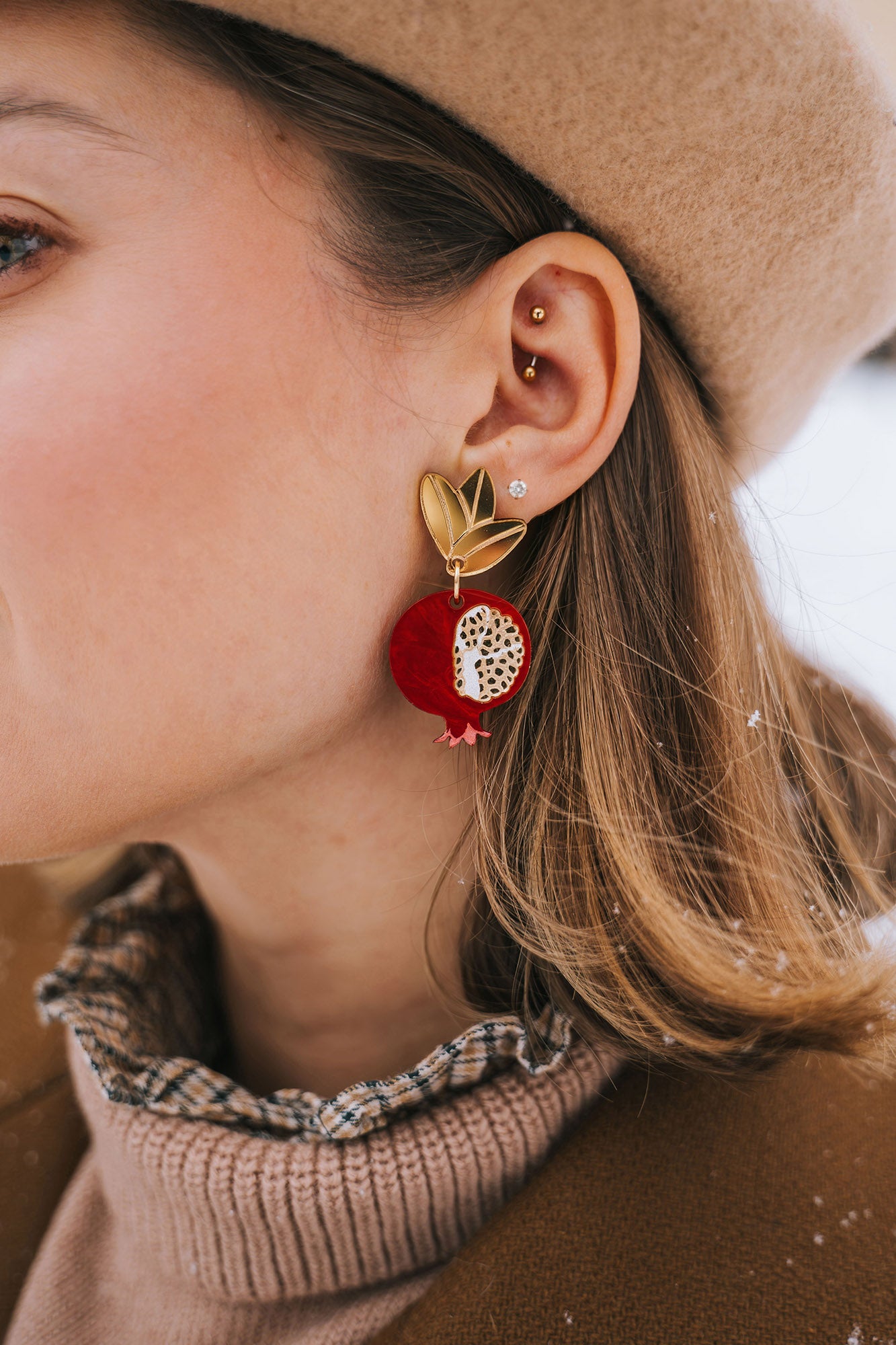 A close-up of a woman's ear wearing a gold stud and a large, handcrafted pomegranate-shaped earring with black leaves. She has a beige beret and a brown coat with a plaid collar. Snowflakes rest on her hair and coat, highlighting bold designs.