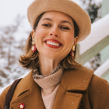 A woman wearing a beige beret, brown coat, and handcrafted earrings with red accents smiles outdoors on a snowy day. Snow is visible on her coat and in the background.