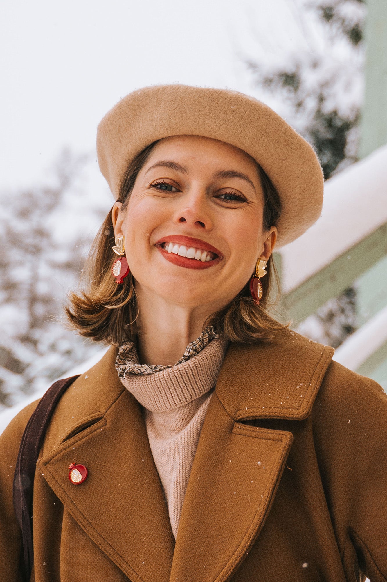 A woman wearing a beige beret, brown coat, and handcrafted earrings with red accents smiles outdoors on a snowy day. Snow is visible on her coat and in the background.