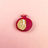 A handcrafted red pomegranate ornament with a gold, textured section showing seeds sits on a light pink background—an elegant fashion accessory for any collection.