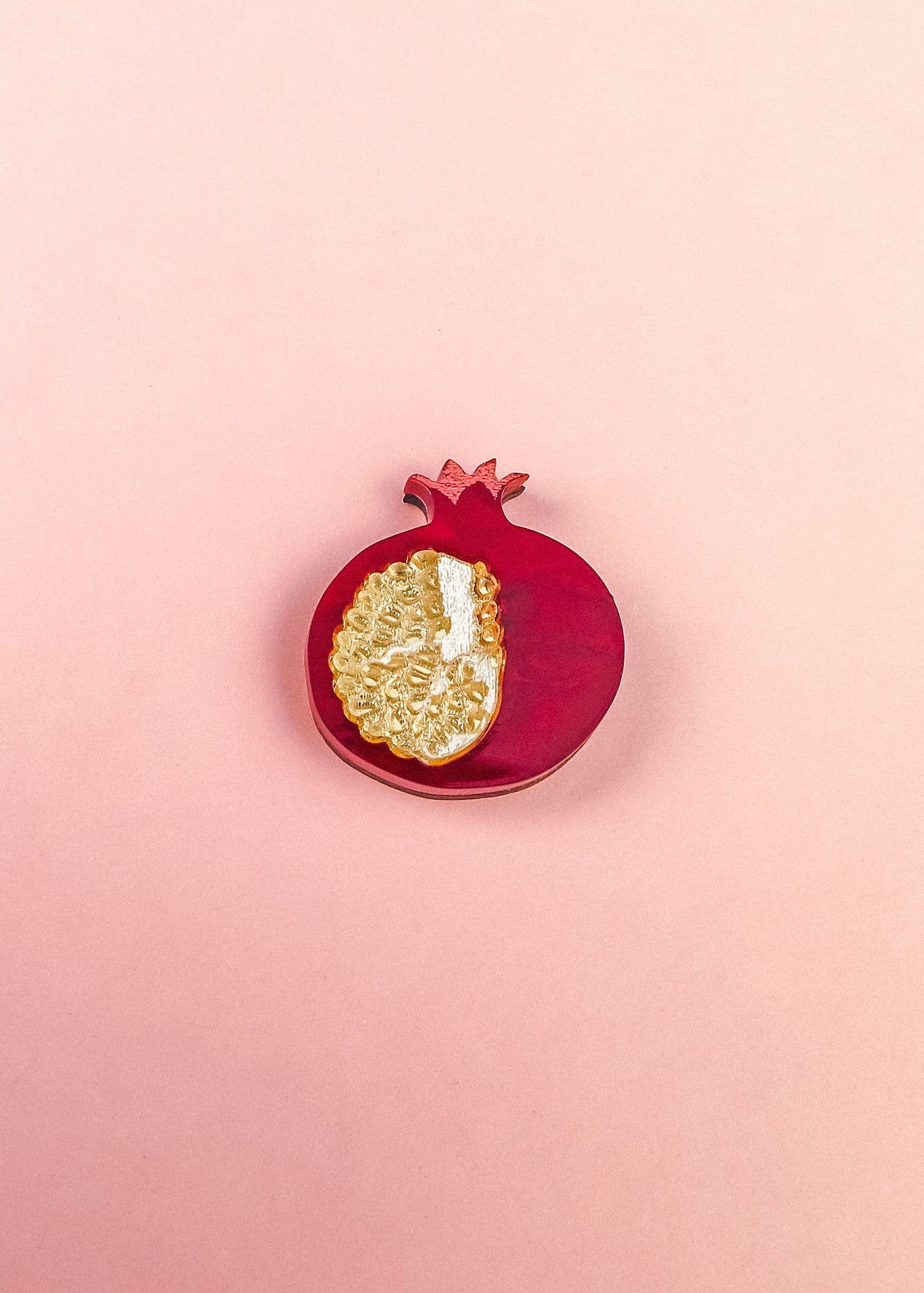 A handcrafted red pomegranate ornament with a gold, textured section showing seeds sits on a light pink background—an elegant fashion accessory for any collection.