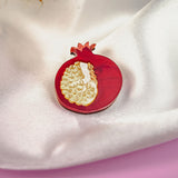 A handcrafted Pomegranate Pin brooch with red and gold details rests on a white satin fabric background, making it an elegant fashion accessory.