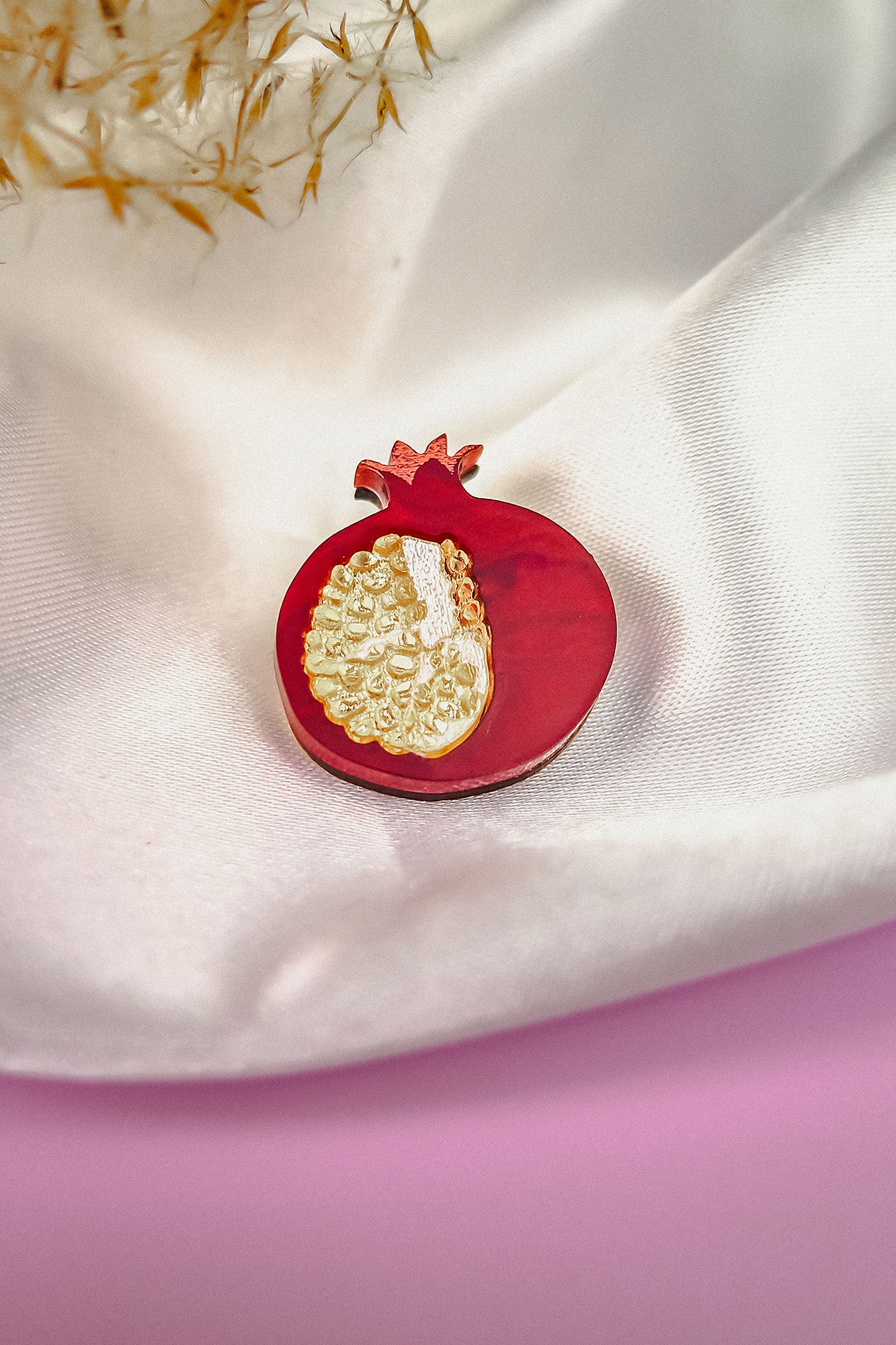 A handcrafted Pomegranate Pin brooch with red and gold details rests on a white satin fabric background, making it an elegant fashion accessory.