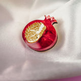 A handcrafted red pomegranate-shaped brooch with gold detailing rests on smooth, white satin fabric, making this Pomegranate Pin a unique fashion accessory. A blurred floral element decorates the upper left corner.