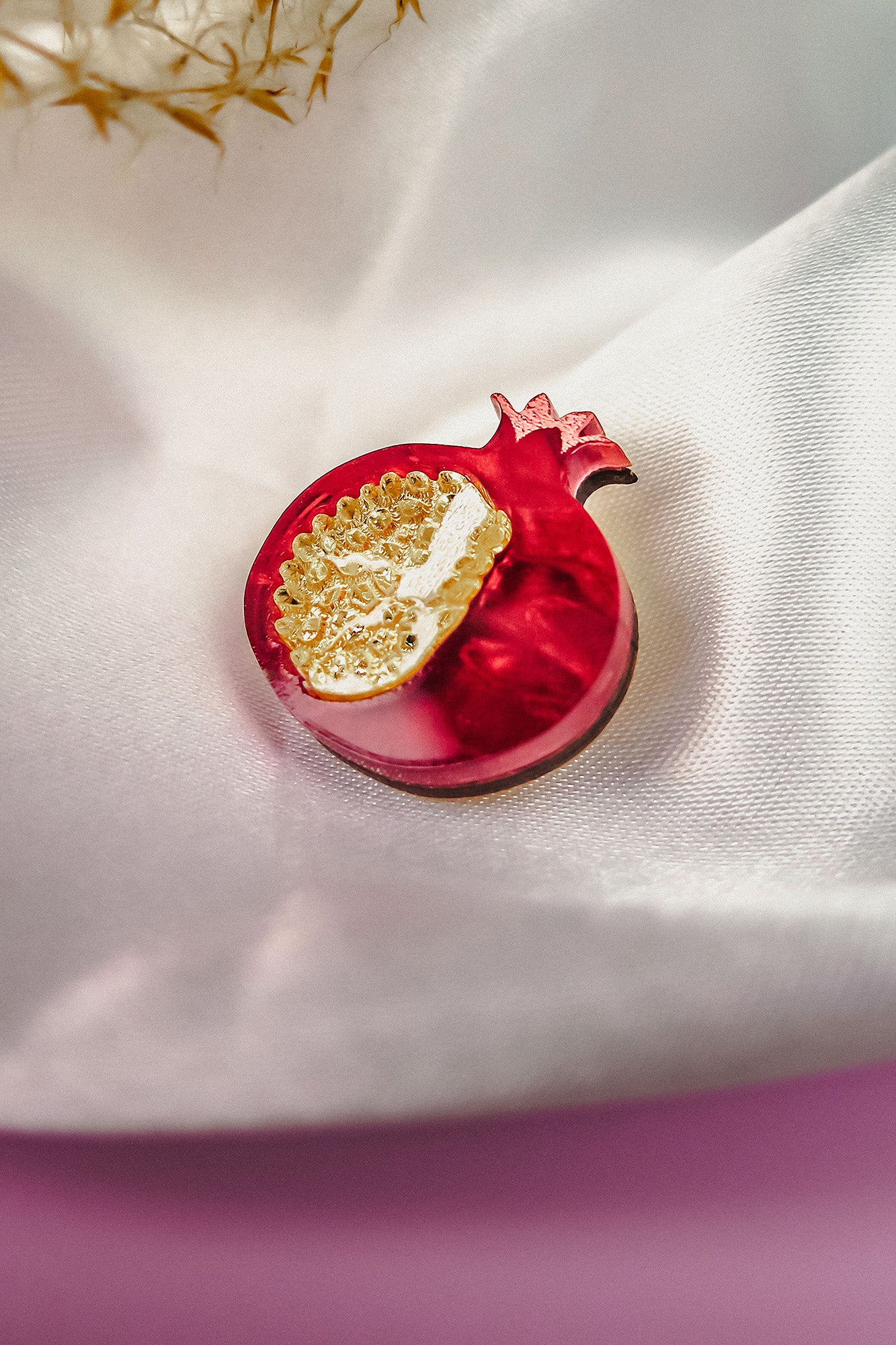 A handcrafted red pomegranate-shaped brooch with gold detailing rests on smooth, white satin fabric, making this Pomegranate Pin a unique fashion accessory. A blurred floral element decorates the upper left corner.