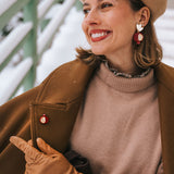 A smiling woman wearing a beige beret, brown coat, tan gloves, and statement earrings with red and gold accents sits outside in a snowy setting, pointing to a handcrafted Pomegranate Pin—a unique fashion accessory on her coat.
