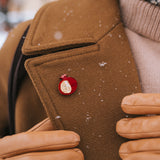 A person wearing a brown coat and tan gloves adjusts the lapel, which is decorated with a handcrafted Pomegranate Pin—a unique fashion accessory. Snowflakes are visible on the coat.