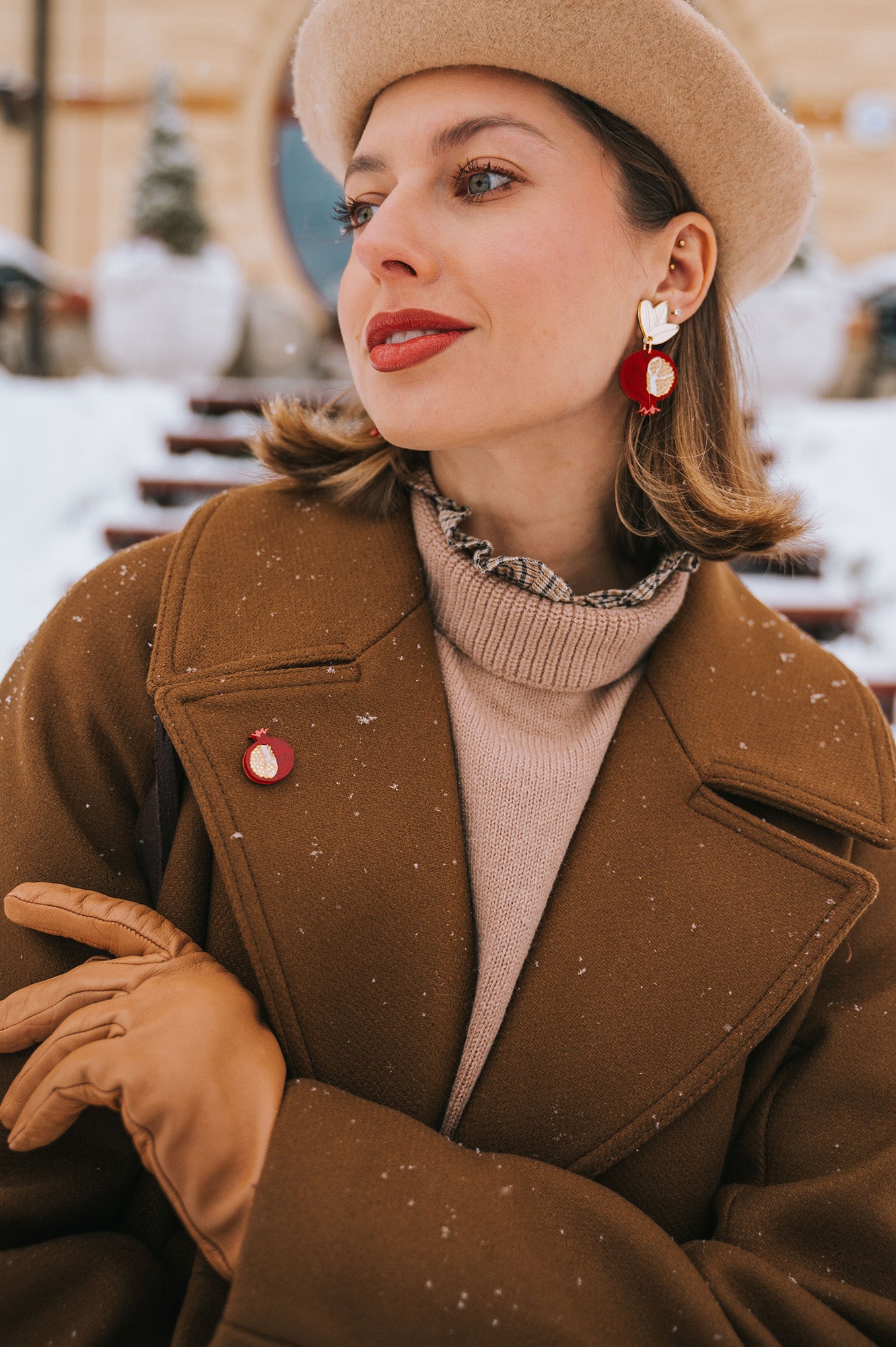 A stylish woman in a beige beret, brown coat, and tan gloves poses outdoors in snowy weather. She wears red and gold handcrafted earrings and a matching Pomegranate Pin fashion accessory, with snowflakes visible on her coat.