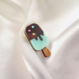 A handcrafted Popsicle Pin shaped like an ice cream bar with a wooden stick, chocolate coating, pastel blue bottom, and three colorful rhinestones sits atop smooth, white satin fabric.