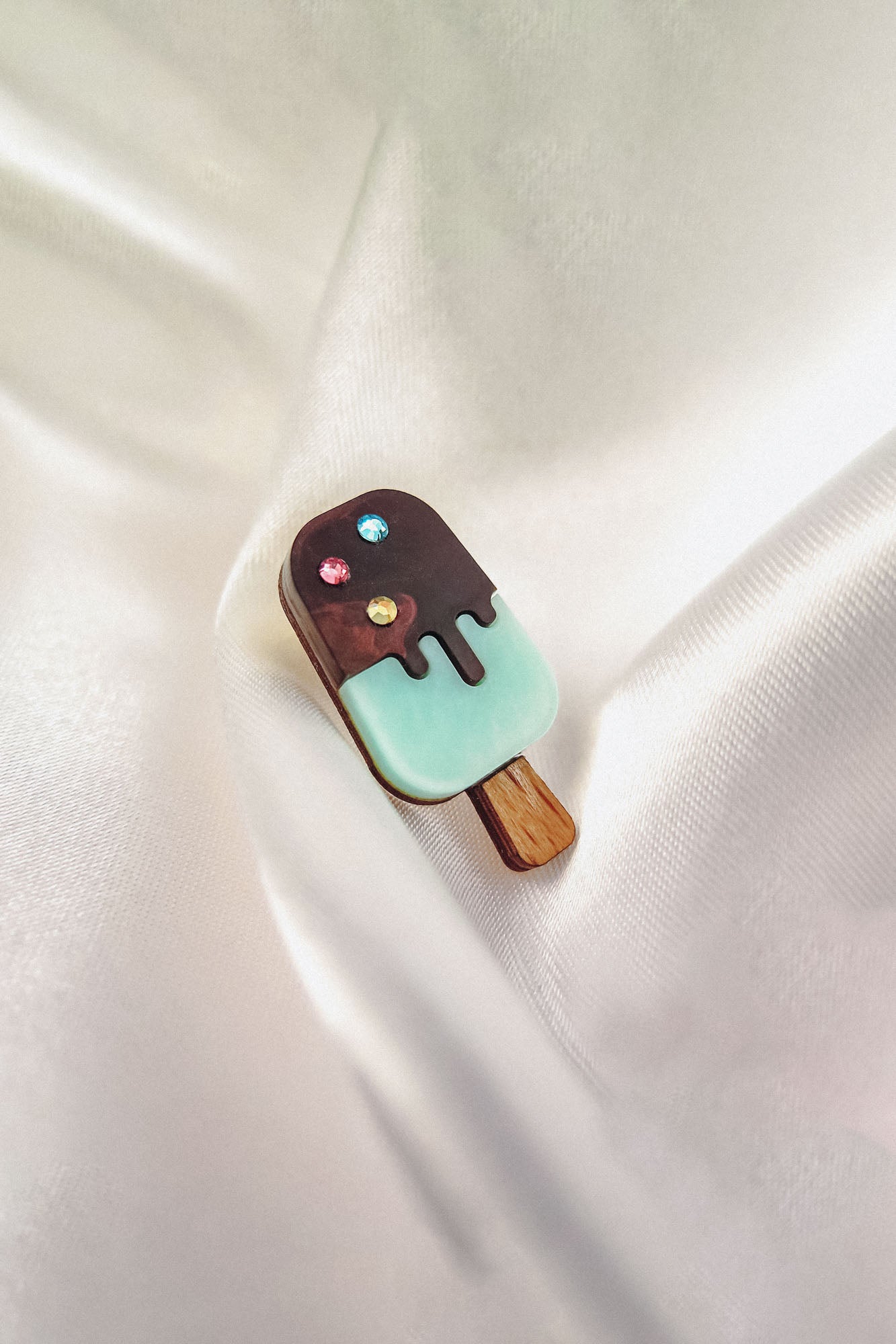 A handcrafted Popsicle Pin shaped like an ice cream bar with a wooden stick, chocolate coating, pastel blue bottom, and three colorful rhinestones sits atop smooth, white satin fabric.