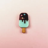 A handcrafted Popsicle Pin shaped like a popsicle with a wooden stick, mint green bottom, chocolate top, and three colored candy decorations, set against a pink and cream gradient background.
