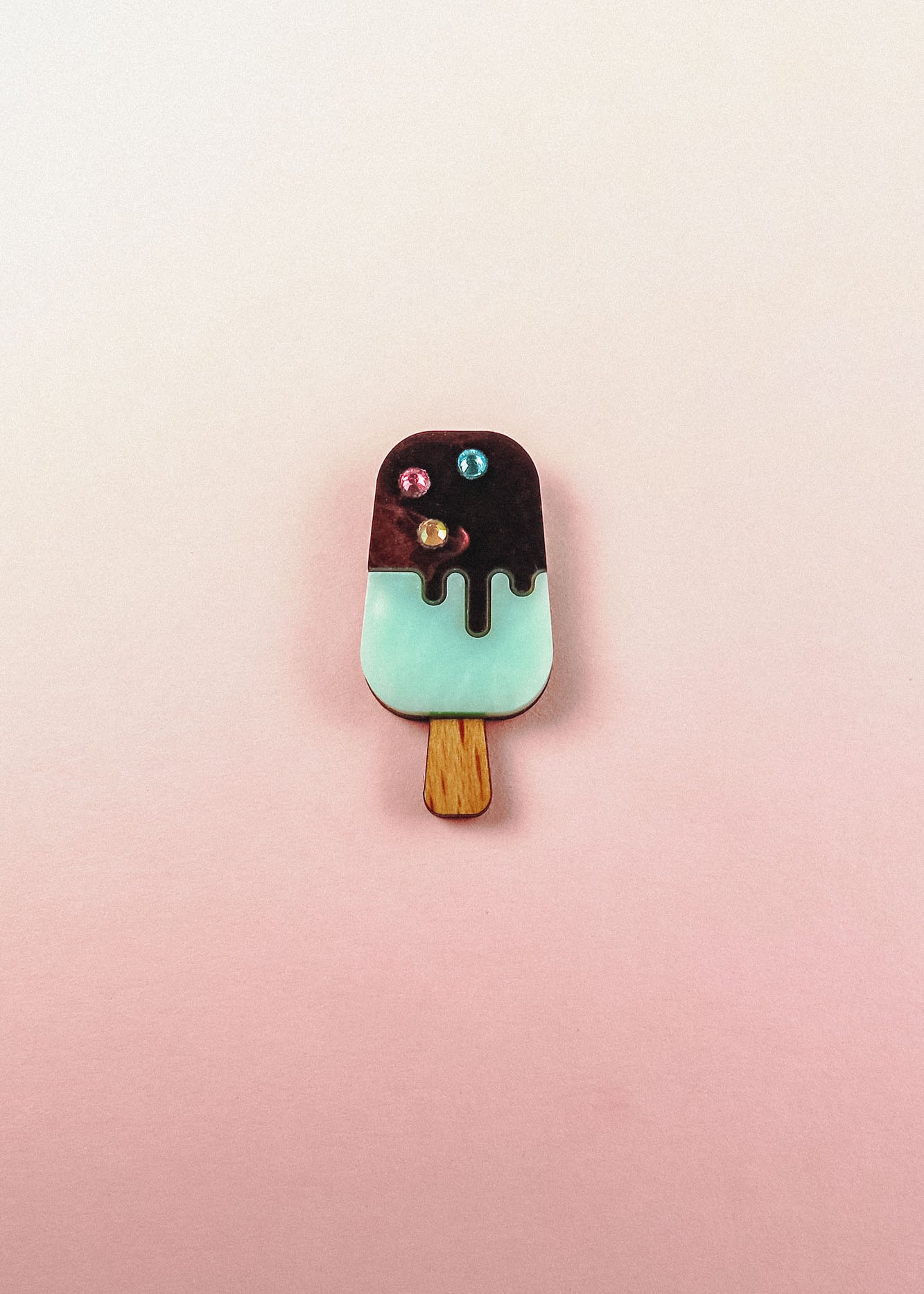 A handcrafted Popsicle Pin shaped like a popsicle with a wooden stick, mint green bottom, chocolate top, and three colored candy decorations, set against a pink and cream gradient background.