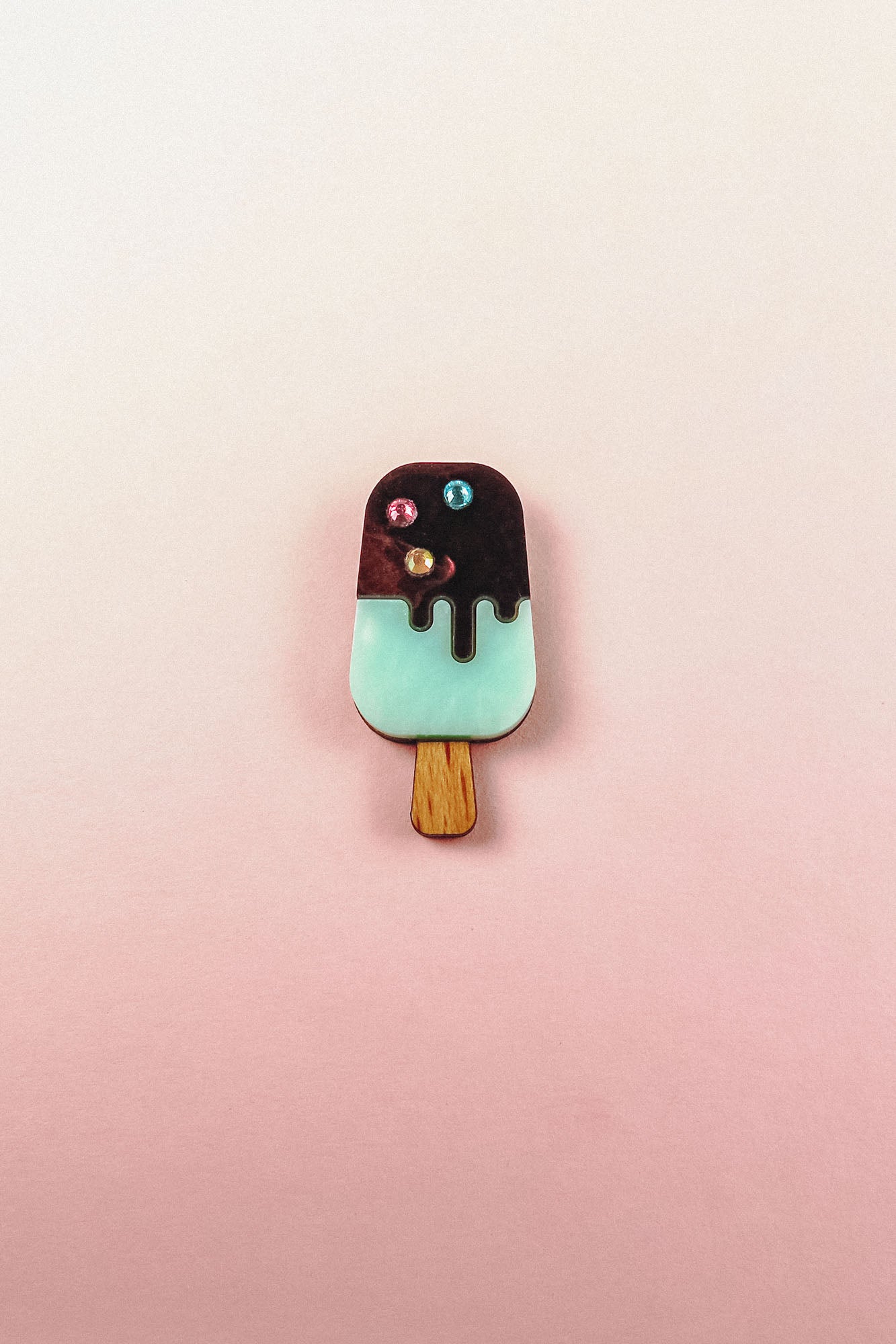 A handcrafted Popsicle Pin shaped like a popsicle with a wooden stick, mint green bottom, chocolate top, and three colored candy decorations, set against a pink and cream gradient background.