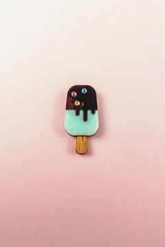 A handcrafted Popsicle Pin shaped like a popsicle with a wooden stick, mint green bottom, chocolate top, and three colored candy decorations, set against a pink and cream gradient background.