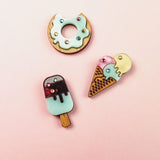 Three cute, colorful enamel pins—including a handcrafted Popsicle Pin, donut, and ice cream cone—sparkle with tiny rhinestones as they’re arranged on a vibrant pink background.