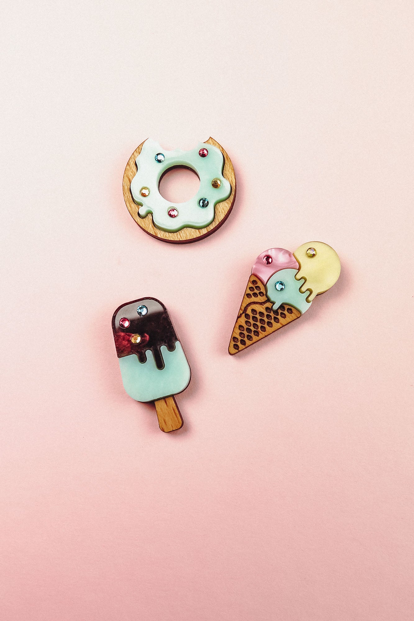 Three cute, colorful enamel pins—including a handcrafted Popsicle Pin, donut, and ice cream cone—sparkle with tiny rhinestones as they’re arranged on a vibrant pink background.