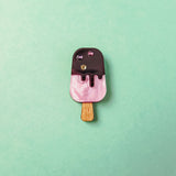 A small, pink and brown handcrafted popsicle pin with glittery gem accents and rhinestones, placed on a mint green background.