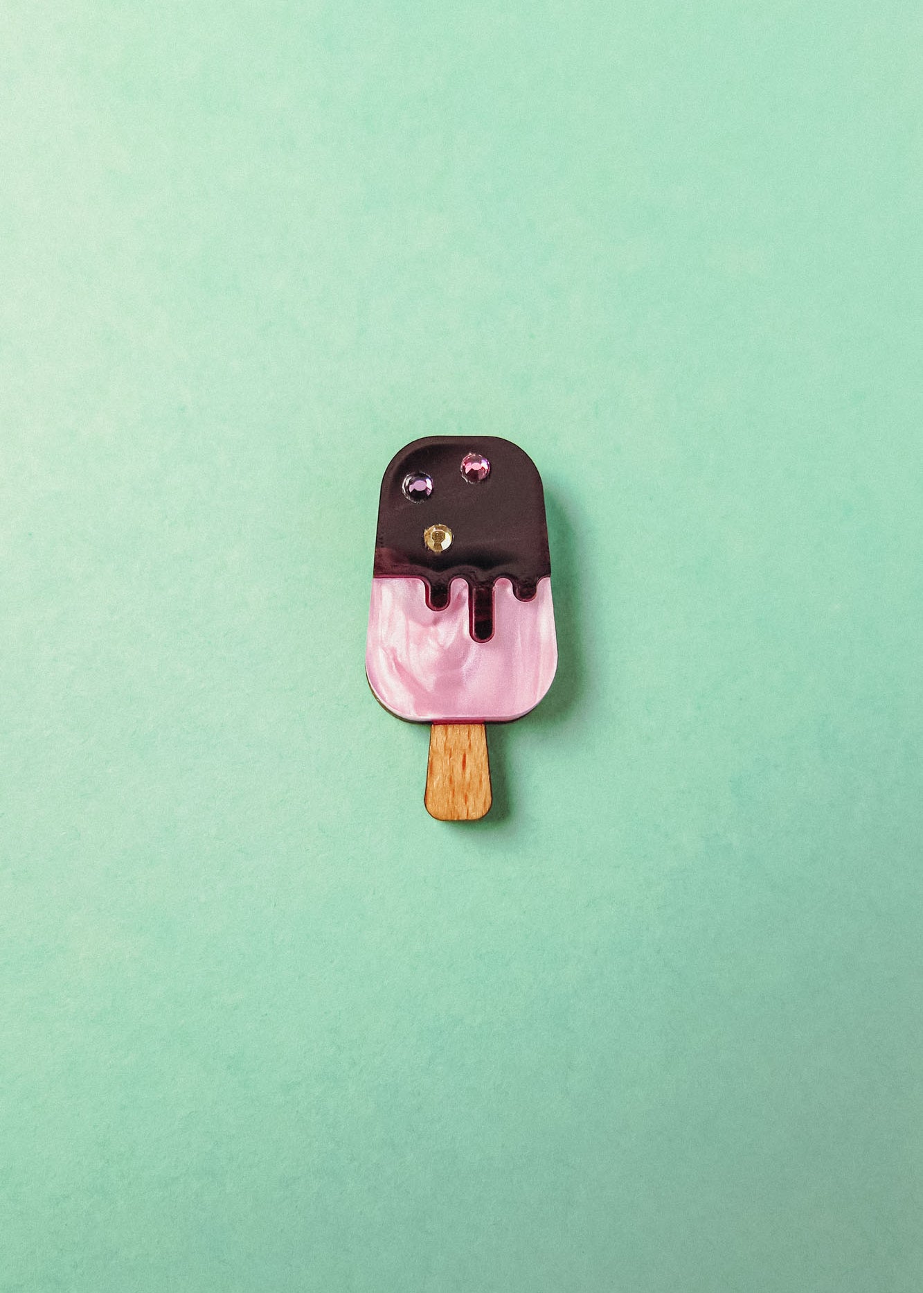 A small, pink and brown handcrafted popsicle pin with glittery gem accents and rhinestones, placed on a mint green background.